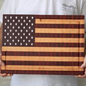 Puede incluir: Una tabla de cortar de madera con forma de bandera estadounidense. La tabla está hecha de madera de arce y nogal y tiene las palabras "We the People" y "Weigelt" grabadas en la parte inferior.