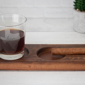 May include: A wooden tray with two circular indentations, one holding a glass of dark liquid with the word "home" etched inside the outline of the state of Texas, and the other holding a brown cigar.