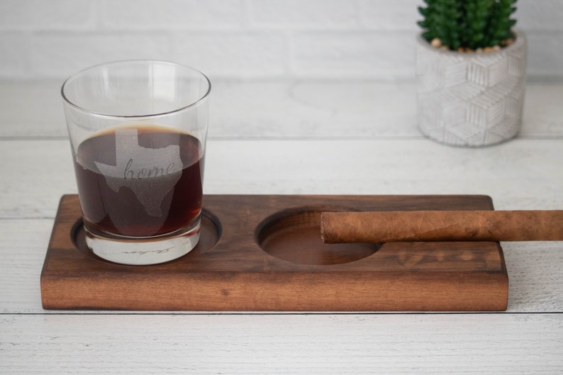May include: A wooden tray with two circular indentations, one holding a glass of dark liquid with the word "home" etched inside the outline of the state of Texas, and the other holding a brown cigar.