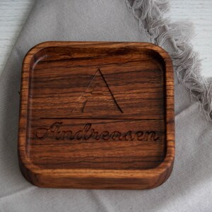 May include: A square wooden tray with a carved triangle and the name "Andreasen" engraved on the surface. The tray is made of dark wood and has a natural finish.