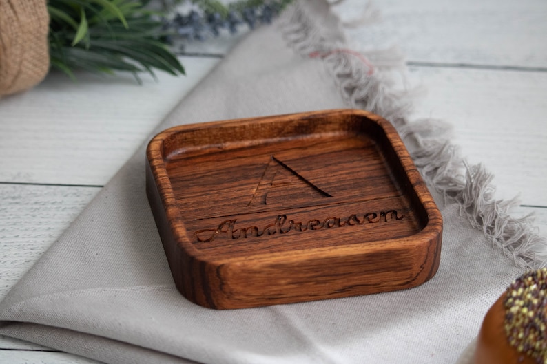 May include: A small, square wooden tray with a dark brown stain. The tray has a triangle and the name "Andreasen" carved into the wood.