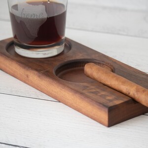 May include: A wooden tray with two circular indentations, one for a glass and one for a cigar. A glass with a dark liquid and the word "home" etched on it sits in one indentation. A brown cigar rests in the other indentation.
