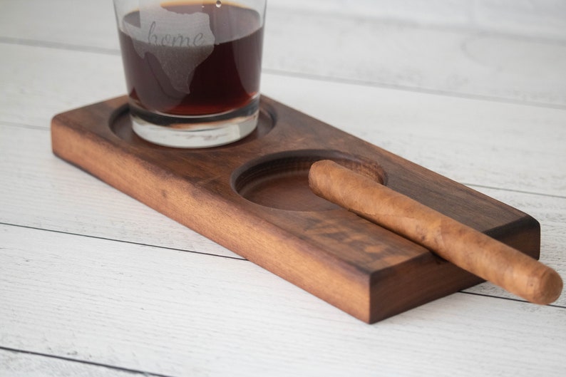 May include: A wooden tray with two circular indentations, one for a glass and one for a cigar. A glass with a dark liquid and the word "home" etched on it sits in one indentation. A brown cigar rests in the other indentation.