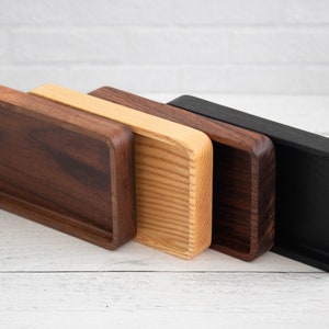 May include: Four rectangular wooden trays in varying shades of brown and black. The trays have rounded corners and a recessed interior. The wood tones range from dark walnut to a natural light wood, and a solid black finish.