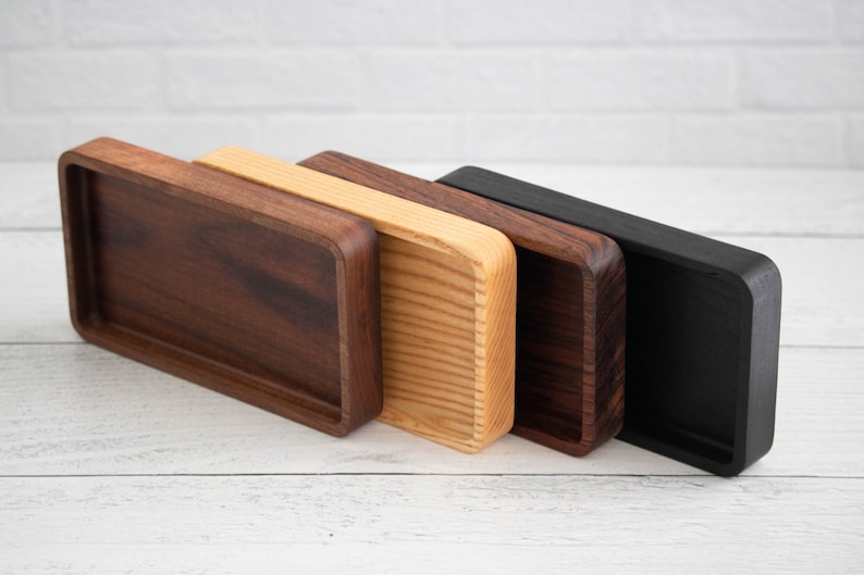 May include: Four rectangular wooden trays in varying shades of brown and black. The trays have rounded corners and a recessed interior. The wood tones range from dark walnut to a natural light wood, and a solid black finish.