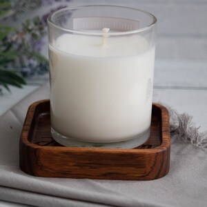 May include: A white candle in a clear glass jar sits on a wooden coaster. The coaster has a dark wood grain and is square shaped.