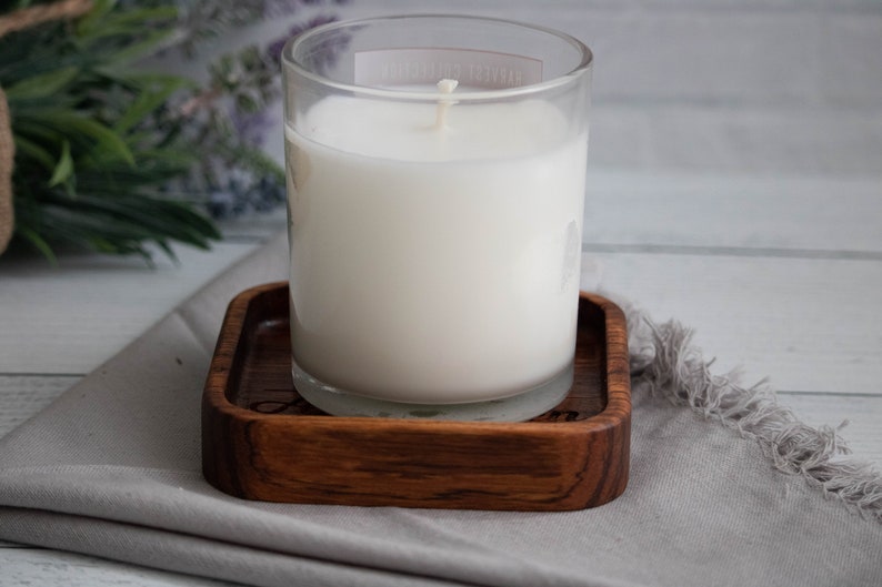 May include: A white candle in a clear glass jar sits on a wooden coaster. The coaster has a dark wood grain and is square shaped.