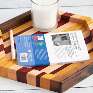 May include: A hexagonal wooden tray constructed from alternating strips of dark walnut, light maple, and reddish padauk wood. The tray holds a white candle in a glass jar, a light-colored pen, and a book titled Scott Thornbury's 101 Grammar Questions.