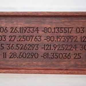 May include: A wooden tray with a dark brown stain. The tray has four lines of text engraved into the wood. The text reads: "06 26.119734 -80.135517 03 27.250763 -80.193992 12 05 36.526293 -121.925224 30 11 28.60290 -81.35036 25"