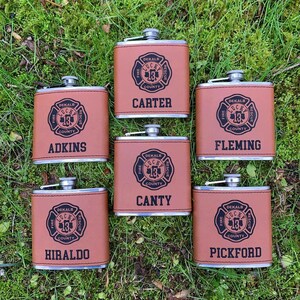 May include: Six brown leather-wrapped flasks with silver tops are arranged on green grass. Each flask features a black graphic of a fire department emblem and a name: Adkins, Carter, Canty, Fleming, Hiraldo, and Pickford. The flasks are for the DeKalb County Fire Rescue.