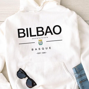 Può includere: Felpa con cappuccio bianca con il testo "BILBAO" in nero, insieme alle coordinate "43°15′25″N 2°55'25"W" e il testo "BASQUE - EST. 1300 -" in nero.