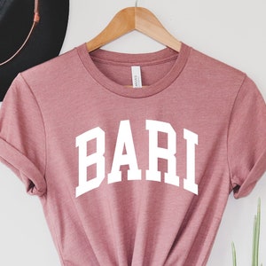 Pode incluir: Uma camiseta cor de rosa pó com a palavra "BARI" impressa em branco na frente. A camiseta está amarrada na parte inferior e está pendurada em um cabide de madeira.