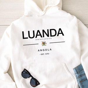 May include: White hooded sweatshirt with black text that reads "LUANDA 8°50'18"S 13°14'4"E ANGOLA - EST. 1576 -" and a small black and white image of a coat of arms.