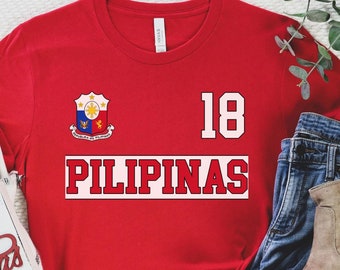 May include: Red t-shirt with a white and blue crest, the text "PILIPINAS" in white with a red outline, and the number "18" in white. The shirt is on a white surface with a white blanket and a pair of blue jeans.