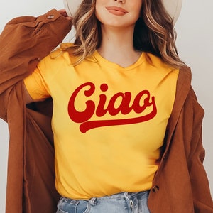 May include: A woman wearing a yellow t-shirt with the word "Ciao" in red cursive lettering. She is also wearing a brown jacket and a white hat.