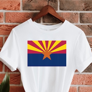 Pode incluir: Camiseta branca com uma imagem impressa da bandeira do estado do Arizona. A bandeira apresenta uma estrela vermelha sobre um fundo azul com raios amarelos e vermelhos que emanam da estrela.