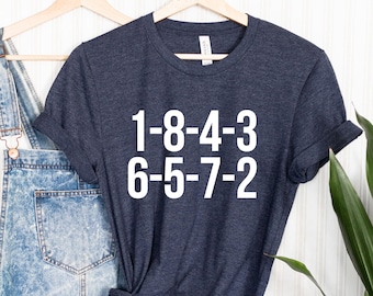 Peut inclure: Un t-shirt bleu foncé avec du texte blanc qui dit "1-8-4-3 6-5-7-2" suspendu à un cintre en bois. Le t-shirt est noué en bas et est affiché sur un fond de bois blanc.