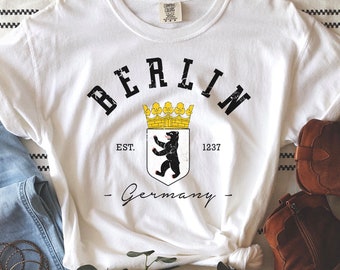 May include: White t-shirt with "BERLIN" in black arched text, a crest with a bear, and "Germany" below. Light blue jeans, brown ankle boots, and gold bracelets are also in the image. The items are arranged on a striped background.