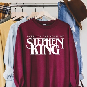 May include: Burgundy sweatshirt with white text that reads "Based on the novel by Stephen King". The sweatshirt is hanging on a rack with other clothing.