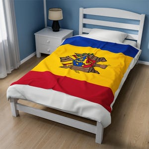 May include: A white twin-sized bed with a yellow, red, and blue blanket featuring the coat of arms of Moldova. The blanket is folded back on the bed.