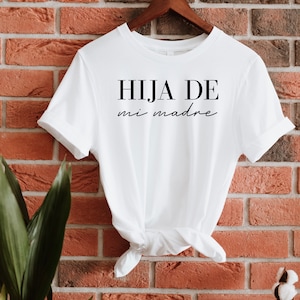 May include: White t-shirt with black text that reads "HIJA DE mi madre" hanging on a wooden hanger against a brick wall.