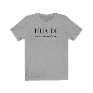 May include: A heather grey t-shirt with the text "HIJA DE mi madre" in black.