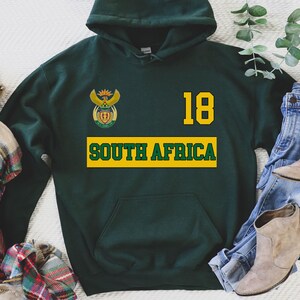 Pu&ograve; includere: Felpa con cappuccio verde con un emblema della bandiera sudafricana, il numero 18 e il testo "SOUTH AFRICA" in giallo.