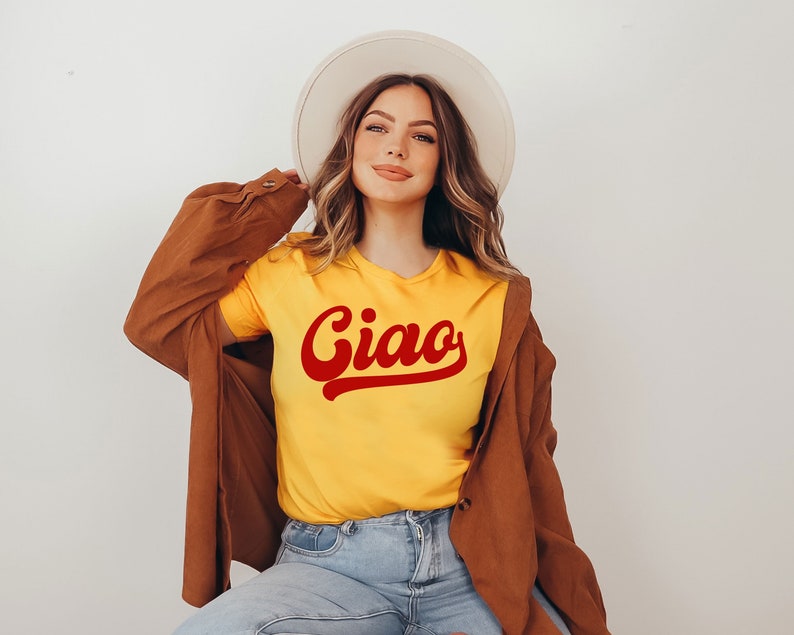 May include: A woman wearing a yellow t-shirt with the word "Ciao" in red cursive lettering. She is also wearing a brown jacket and a white hat.