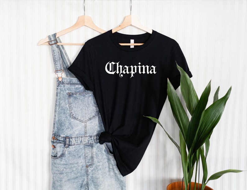 May include: Black t-shirt with white text that reads "Chapina" in a gothic font. The shirt is hanging on a wooden hanger and is styled with denim overalls.