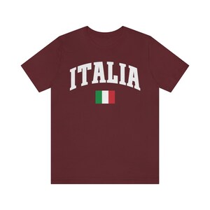 Pu&ograve; includere: Una maglietta color granata con la scritta "ITALIA" in bianco in un font ad arco. La bandiera italiana &egrave; sotto il testo.