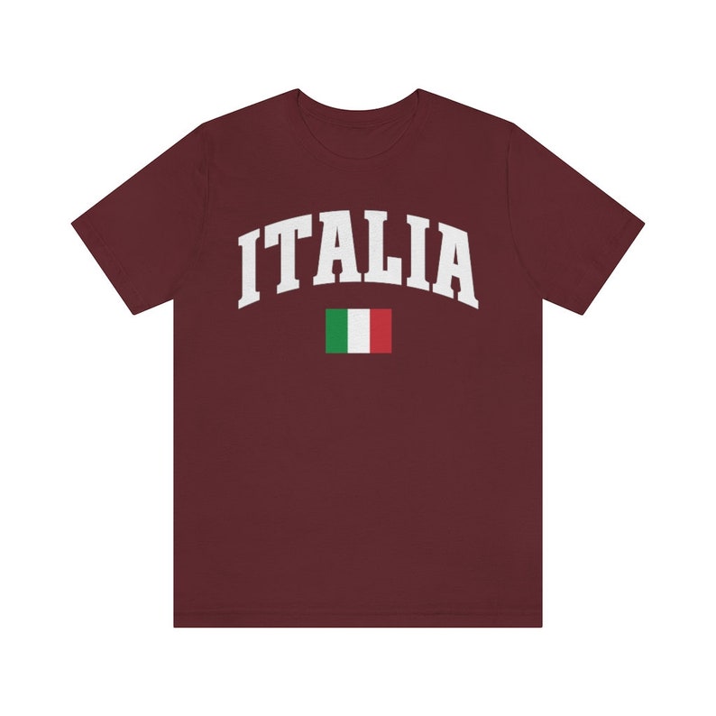 Pu&ograve; includere: Una maglietta color granata con la scritta "ITALIA" in bianco in un font ad arco. La bandiera italiana &egrave; sotto il testo.