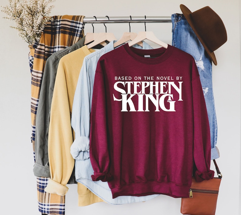 May include: Burgundy sweatshirt with white text that reads "Based on the novel by Stephen King". The sweatshirt is hanging on a rack with other clothing.