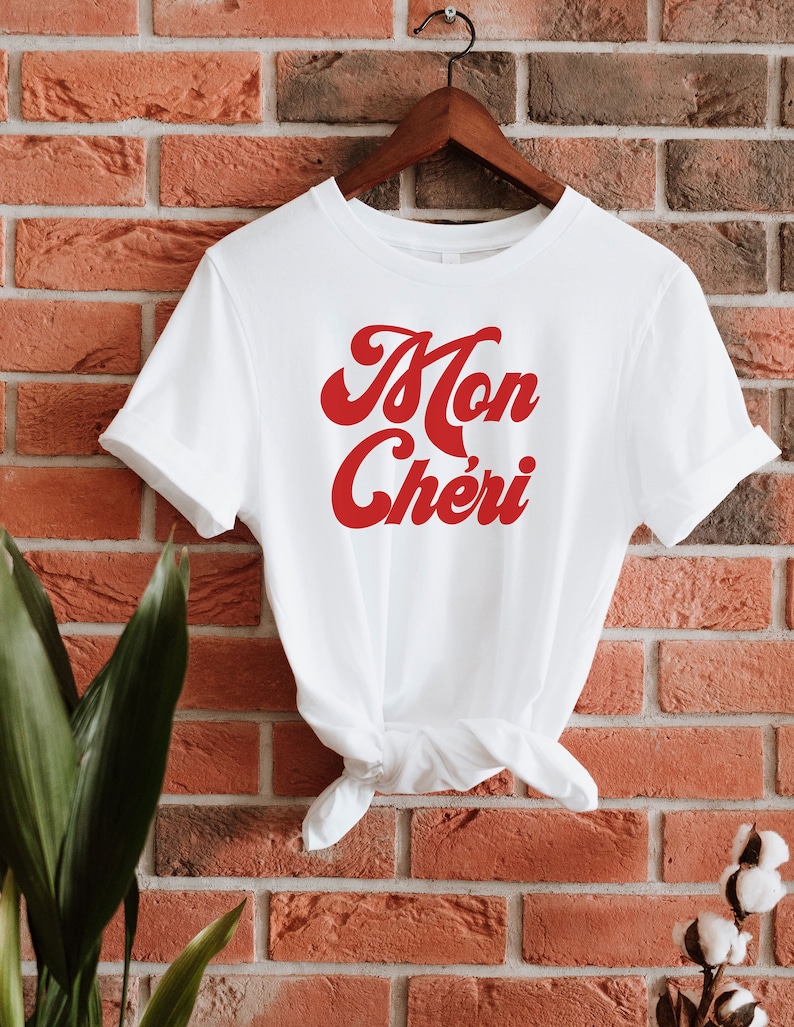 May include: White t-shirt with a red retro-style graphic that says "Mon Ch&eacute;ri" hanging on a wooden hanger against a brick wall.