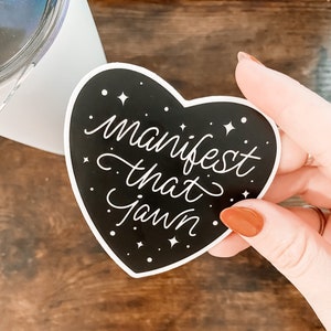 Pu&ograve; includere: Un adesivo nero a forma di cuore con testo bianco che recita "manifest that jawnn" e stelle bianche su sfondo nero.