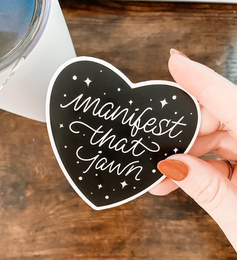 Pu&ograve; includere: Un adesivo nero a forma di cuore con testo bianco che recita "manifest that jawnn" e stelle bianche su sfondo nero.