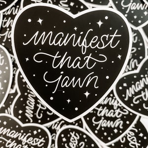 Pu&ograve; includere: Un adesivo nero a forma di cuore con testo bianco che recita "manifest that jawn". L'adesivo &egrave; circondato da altri adesivi simili con lo stesso testo.