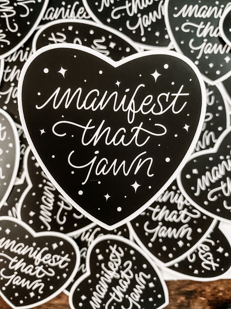 Pu&ograve; includere: Un adesivo nero a forma di cuore con testo bianco che recita "manifest that jawn". L'adesivo &egrave; circondato da altri adesivi simili con lo stesso testo.