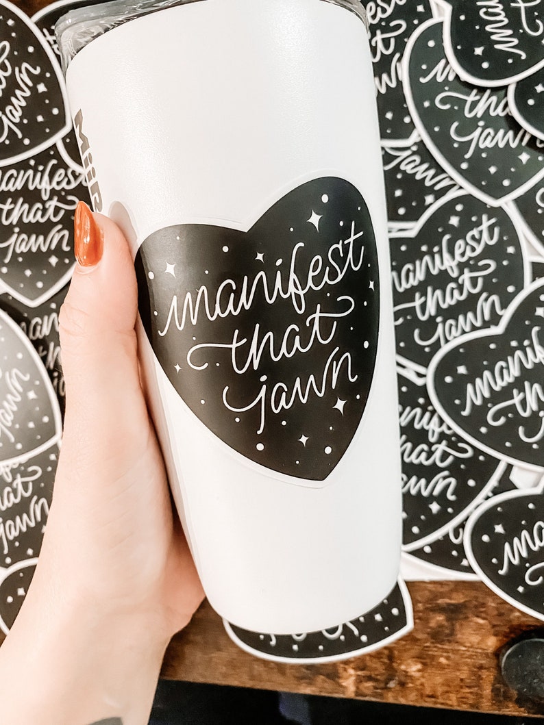 Pu&ograve; includere: Un bicchiere bianco con un adesivo nero a forma di cuore che dice "manifest that jawn" in lettere bianche in corsivo. L'adesivo ha uno sfondo nero con stelle bianche.