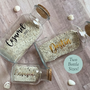 May include: Three glass bottles with cork stoppers filled with sand. The bottles are labeled with the names of vacation destinations: Cozumel 2021, Destin FL 2021, and Engaged. Two bottle sizes are shown.