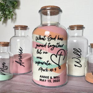 May include: A clear glass bottle filled with layered pink, orange, and mint sand, featuring the black text What God has joined together let no one separate. Mark 10:9, a heart with a cross, and the names Annie & Will with date May 15, 2025.