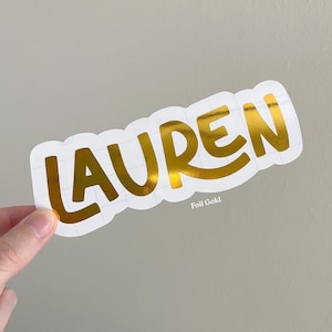 May include: A die-cut sticker featuring the name LAUREN in a bold, metallic gold foil finish. The letters are set against a white backing with a grid pattern visible, and the text Foil Gold is printed in small white letters below the name.