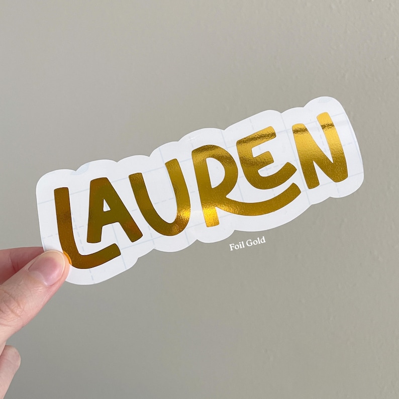 May include: A die-cut sticker featuring the name LAUREN in a bold, metallic gold foil finish. The letters are set against a white backing with a grid pattern visible, and the text Foil Gold is printed in small white letters below the name.