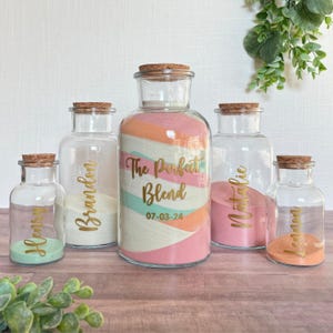 May include: Five clear glass jars with cork stoppers, filled with layers of colored sand. The central large jar features the gold text "The Perfect Blend" and "07-03-24". Four smaller jars are labeled with the names "Henry", "Brandon", "Natalie", and "Lemon".