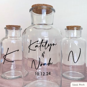 Op de afbeelding: Drie doorzichtige glazen potten met kurken stoppen. De middelste pot heeft zwarte vinyltekst met Katelyn & Noah en de datum 10.12.24. De linkerpot toont een zwarte letter K en de rechterpot toont een zwarte letter N.