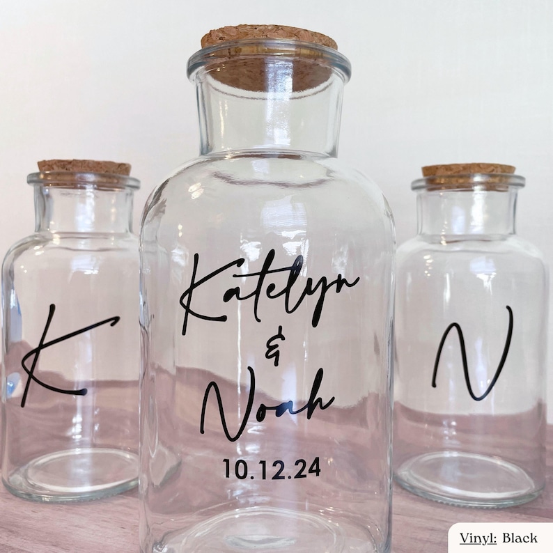Op de afbeelding: Drie doorzichtige glazen potten met kurken stoppen. De middelste pot heeft zwarte vinyltekst met Katelyn & Noah en de datum 10.12.24. De linkerpot toont een zwarte letter K en de rechterpot toont een zwarte letter N.