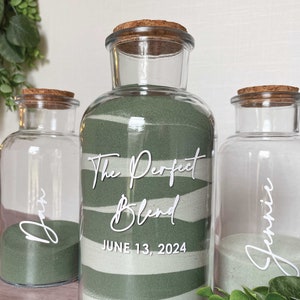 May include: Three clear glass bottles with cork stoppers. The center bottle has green and white sand with the text "The Perfect Blend" and "June 13, 2024". The other two bottles have green sand and the names "Dan" and "Jennie".
