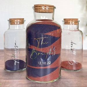 May include: Three glass sand ceremony vases with cork stoppers. The center vase is filled with blue and red sand and has the text "The Brights" and the date "12.10.22". The other two vases have names in silver script and are filled with sand.