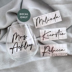 May include: Three clear vinyl stickers with black script lettering. The stickers read "Melinda", "Kieren", and "Rebecca". A clear glass wine glass with a black script decal that reads "Mrs. Ackley".