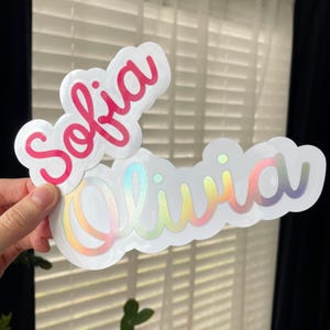 May include: Two custom die-cut stickers with white borders. The top sticker features the name Sofia in a bright pink script font. The bottom sticker displays the name Olivia in a holographic, iridescent script font that shifts colors.