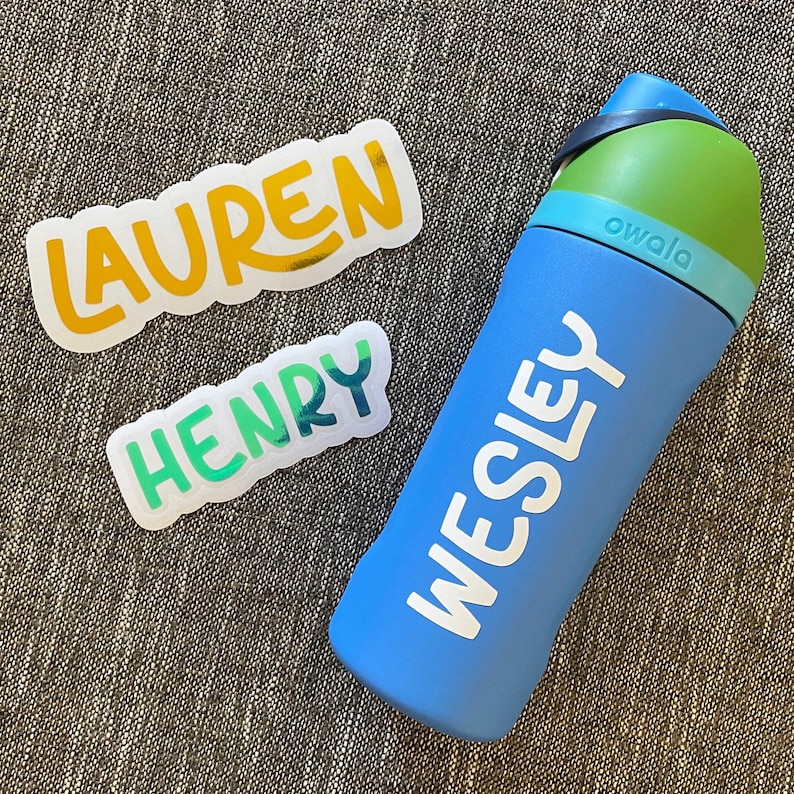 May include: A blue Owala water bottle with the name WESLEY printed in white. Beside the bottle are two die-cut vinyl stickers: one with the name LAUREN in yellow and one with the name HENRY in a green to blue gradient, both with white borders.